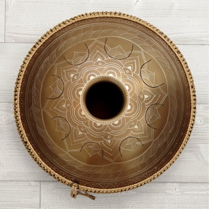 Puede incluir: Un handpan de color dorado con un diseño de mandala y una correa de cuero marrón. El handpan es un instrumento musical que se toca golpeando la superficie.