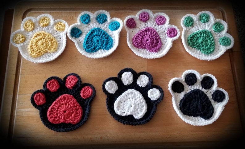 May include: Seven crocheted paw print coasters in various colors, including yellow, teal, purple, green, red, white, and black.