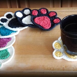 May include: A set of crocheted paw print coasters in various colors, including white, black, teal, purple, green, and red. A black mug is sitting on a white paw print coaster with yellow accents.