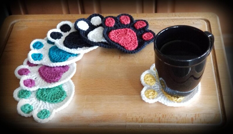 May include: A set of crocheted paw print coasters in various colors, including white, black, teal, purple, green, and red. A black mug is sitting on a white paw print coaster with yellow accents.