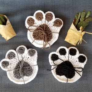 May include: Three crocheted paw print coasters. One is brown, one is gray, and one is black. Each coaster is tied with a string.