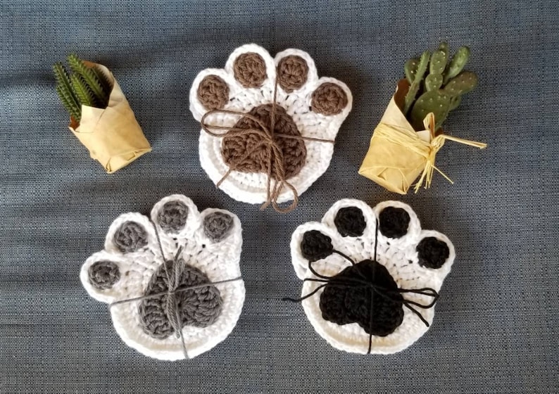 May include: Three crocheted paw print coasters in brown, gray, and black. Each coaster is made of white yarn with a contrasting colored paw print in the center.