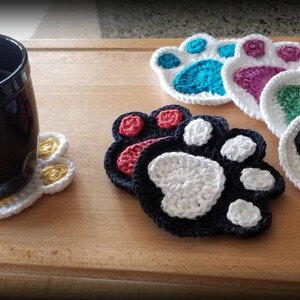 May include: Five crocheted paw print coasters in various colors, including teal, purple, green, red, and black. The coasters are on a wooden surface with a black mug on a white paw print coaster.