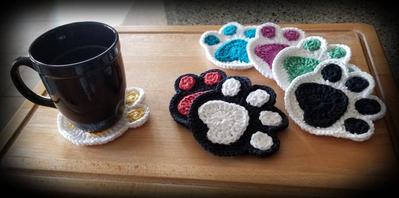 May include: Five crocheted paw print coasters in various colors, including teal, purple, green, red, and black. The coasters are on a wooden surface with a black mug on a white paw print coaster.