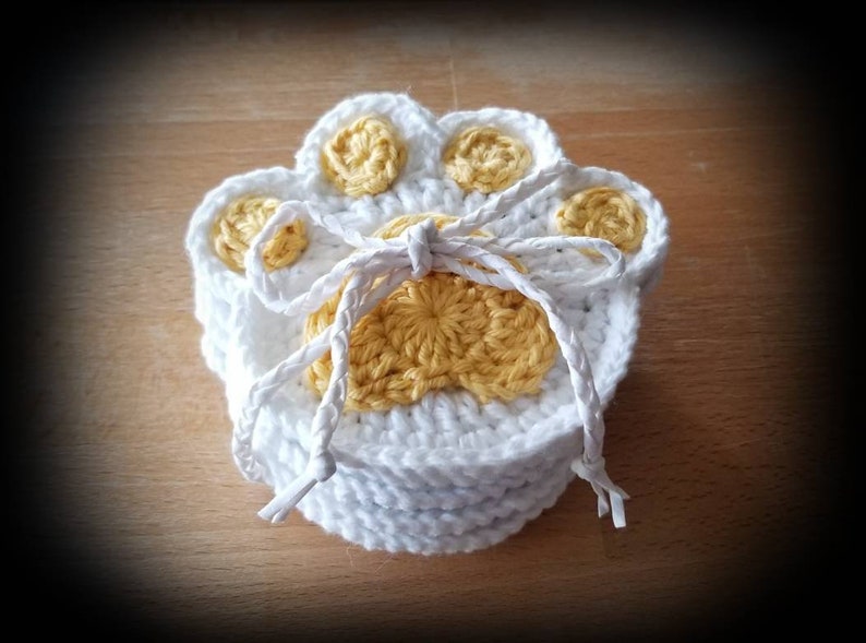 May include: Set of four crocheted white paw print coasters with yellow centers. The coasters are tied together with a white string.