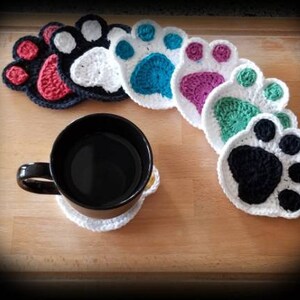 May include: Six crocheted paw print coasters in various colors, including red, white, blue, purple, green, and black. The coasters are arranged in a semi-circle on a wooden surface. A black coffee mug is sitting on one of the coasters.