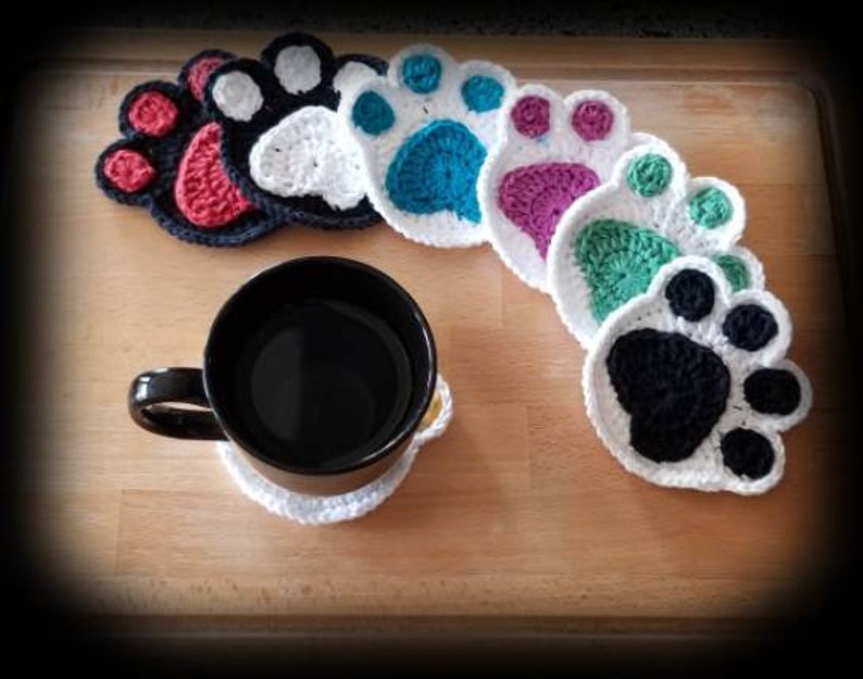 May include: Six crocheted paw print coasters in various colors, including red, white, blue, purple, green, and black. The coasters are arranged in a semi-circle on a wooden surface. A black coffee mug is sitting on one of the coasters.