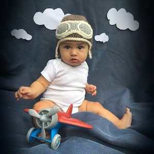 May include: A baby wearing a brown and grey aviator hat with goggles sits on a blue blanket holding a red and blue toy aeroplane.