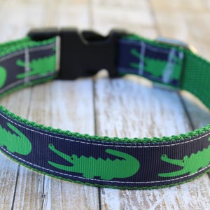 May include: A pet collar featuring a repeating pattern of bright green alligators on a dark navy blue background. The collar is constructed with a durable green webbing base, a black plastic side-release buckle, and a metal D-ring for attachment.