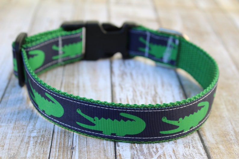 May include: A pet collar featuring a repeating pattern of bright green alligators on a dark navy blue background. The collar is constructed with a durable green webbing base, a black plastic side-release buckle, and a metal D-ring for attachment.