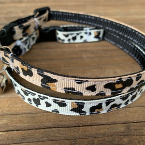 Puede incluir: Un collar de gato de estampado de leopardo blanco y negro con una campanilla. El collar est&aacute; hecho de dos capas de tela, una con fondo blanco y la otra con fondo beige.