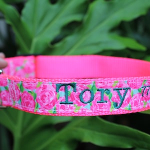 May include: A neon pink nylon pet collar featuring a floral pattern of pink roses and green leaves. The collar includes a silver metal D-ring, a black plastic buckle, and the name Tory embroidered in dark green thread on the front surface.