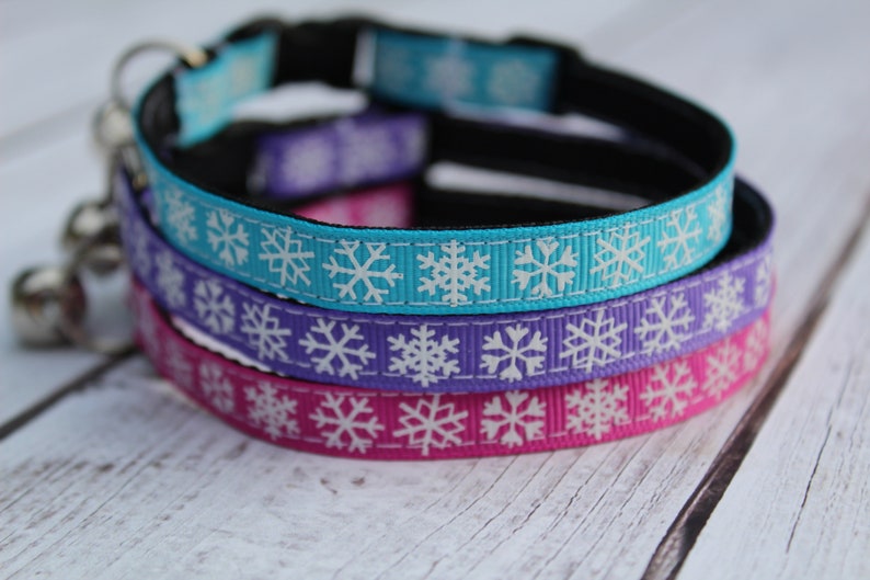 May include: Three cat collars with snowflake patterns. The collars are turquoise, purple, and pink with white snowflakes. The collars are made of ribbon and have a black adjustable strap.