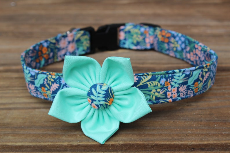 K&ouml;nnte beinhalten: Ein Haustierhalsband mit einer dunkelblauen Stoffbasis und einem mehrfarbigen Blumenmuster in Rosa-, Orange- und Gr&uuml;nt&ouml;nen. Eine gro&szlig;e, einfarbige mintgr&uuml;ne Stoffblume mit einem passenden, blumig gemusterten Knopf in der Mitte ist vorne befestigt.
