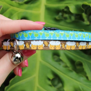Op de afbeelding: Een blauwe en bruine kattenhalsband met een bel. De halsband is versierd met een patroon van groene cocktailglazen en bruine bierpullen.