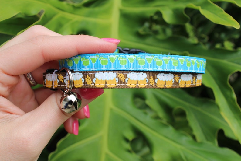 Op de afbeelding: Een blauwe en bruine kattenhalsband met een bel. De halsband is versierd met een patroon van groene cocktailglazen en bruine bierpullen.