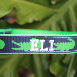 May include: A bright green and navy blue pet collar featuring a repeating pattern of green alligators. The collar includes a silver metal D-ring and is personalized with the name ELI embroidered in white capital letters on the navy ribbon.
