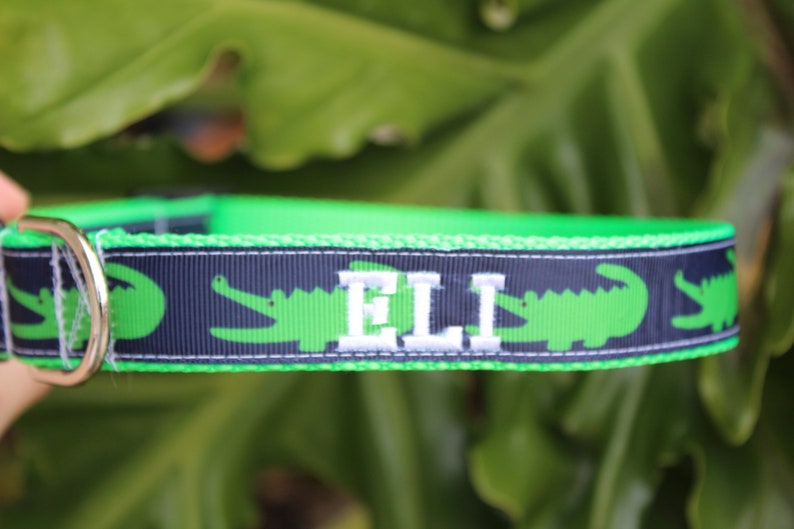 May include: A bright green and navy blue pet collar featuring a repeating pattern of green alligators. The collar includes a silver metal D-ring and is personalized with the name ELI embroidered in white capital letters on the navy ribbon.