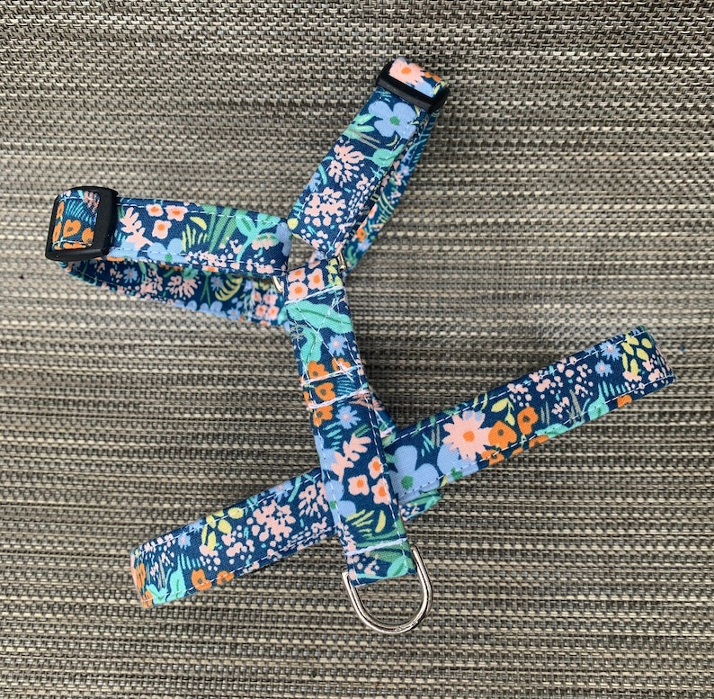 K&ouml;nnte beinhalten: Ein Haustiergeschirr mit dunkelblauem Hintergrund und einem lebendigen Blumenmuster in Hellblau, Orange und Rosa. Das Geschirr verf&uuml;gt &uuml;ber verstellbare schwarze Kunststoffschnallen, wei&szlig;e N&auml;hte und einen silbernen Metall-D-Ring f&uuml;r die Leine.