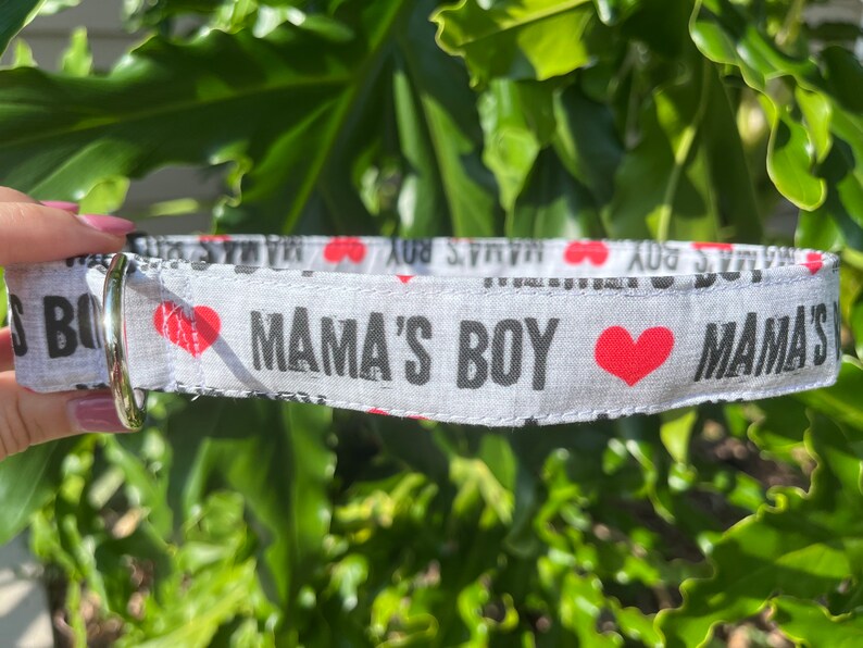 May include: A light gray fabric pet collar featuring the repeating black text MAMA'S BOY and small red heart icons. The collar includes a silver-toned metal D-ring attached to one end for leash connection, showcasing a clear, repetitive print design.