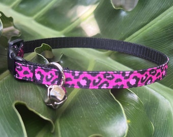 May include: A pink and black leopard print cat collar with a silver bell.
