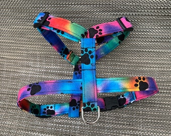 May include: A pet harness featuring a vibrant rainbow tie-dye pattern with black paw print graphics. The harness includes adjustable fabric straps, black plastic side-release buckles, and a silver-toned metal D-ring for leash attachment.
