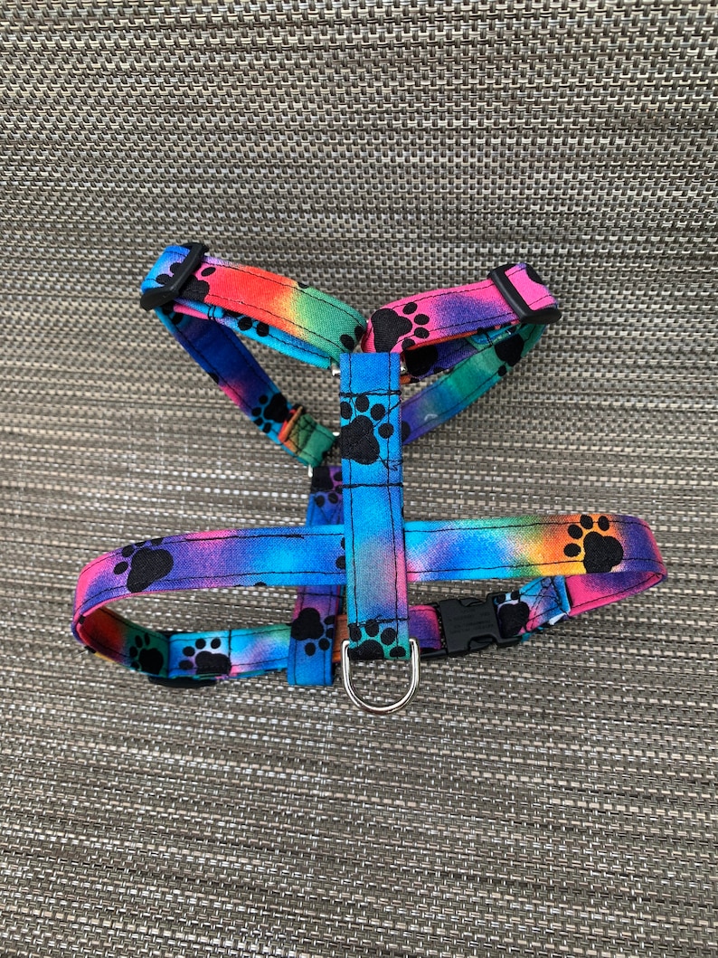 May include: A pet harness featuring a vibrant rainbow tie-dye pattern with black paw print graphics. The harness includes adjustable fabric straps, black plastic side-release buckles, and a silver-toned metal D-ring for leash attachment.