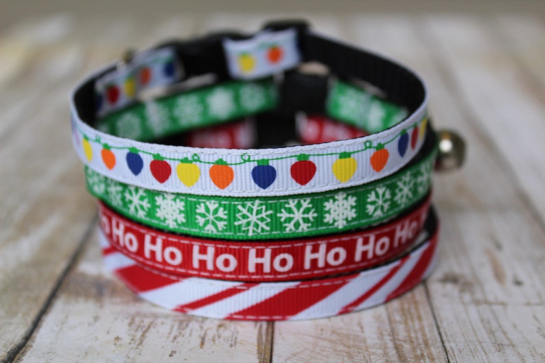 May include: A stack of four festive cat collars with Christmas themes. The collars feature red and white candy cane stripes, a red and white "Ho Ho Ho" design, a green and white snowflake pattern, and a white collar with a string of colorful lights.