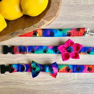 May include: A set of pet accessories featuring a tie-dye pattern with black paw prints. The set includes a leash with a metal clip, a collar with a large pink fabric flower attachment, and a collar with a matching tie-dye fabric bow tie.