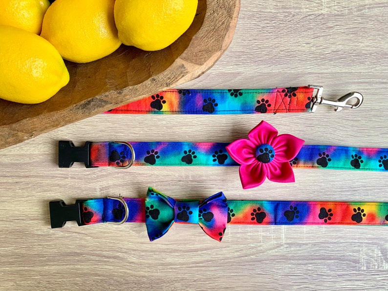 May include: A set of pet accessories featuring a tie-dye pattern with black paw prints. The set includes a leash with a metal clip, a collar with a large pink fabric flower attachment, and a collar with a matching tie-dye fabric bow tie.