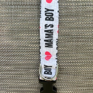 May include: A light gray fabric pet collar featuring a repeating pattern of the black text MAMA'S BOY and red heart icons. The collar includes a silver metal D-ring for leash attachment and a black plastic side-release buckle for secure fastening.