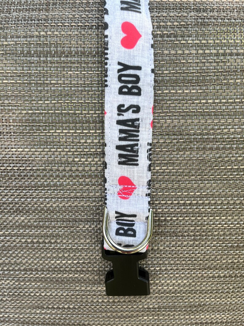 May include: A light gray fabric pet collar featuring a repeating pattern of the black text MAMA'S BOY and red heart icons. The collar includes a silver metal D-ring for leash attachment and a black plastic side-release buckle for secure fastening.