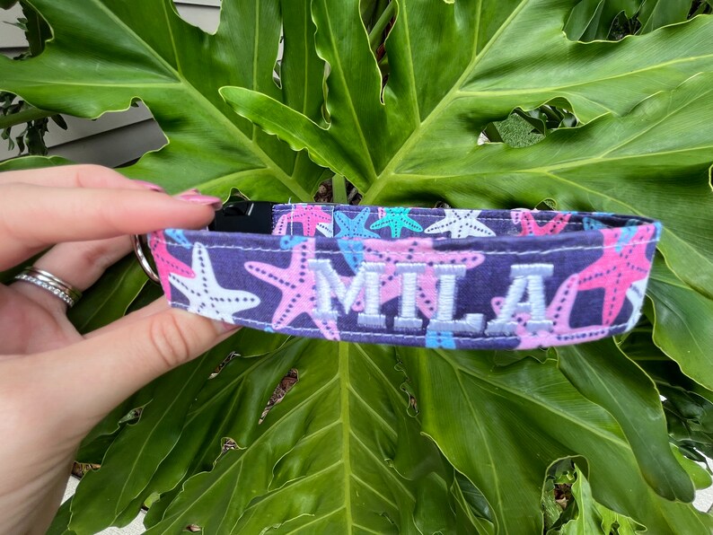 May include: A dark blue dog collar with a pink and white starfish pattern and the name "Mila" embroidered in white.