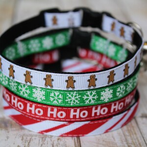 May include: A stack of four festive cat collars with Christmas themes. The top collar features gingerbread men, the second has snowflakes, the third says "Ho Ho Ho", and the bottom has red and white candy cane stripes.