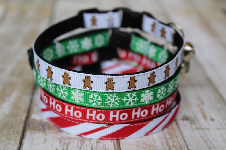 May include: A stack of four festive cat collars with Christmas themes. The top collar features gingerbread men, the second has snowflakes, the third says "Ho Ho Ho", and the bottom has red and white candy cane stripes.