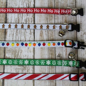 May include: Five different Christmas themed cat collars with bells. The collars are red with white "Ho Ho Ho" text, white with gingerbread men, white with colorful lights, green with white snowflakes, and red and white striped.