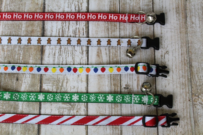 May include: Five different Christmas themed cat collars with bells. The collars are red with white "Ho Ho Ho" text, white with gingerbread men, white with colorful lights, green with white snowflakes, and red and white striped.