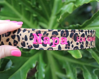 May include: A pet collar featuring a tan and black leopard print pattern. The collar is embroidered with the name Napa in bright pink thread, followed by the phone number 504-439-6000 in the same pink embroidery, stitched horizontally across the fabric.