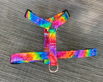 May include: A colorful dog harness with a tie-dye pattern in shades of red, orange, yellow, green, blue, and purple. The harness has a D-ring for leash attachment and adjustable straps. The harness is designed for small to medium-sized dogs.