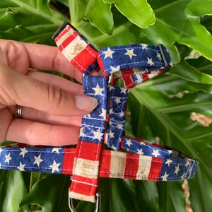 Könnte beinhalten: Ein Haustiergeschirr mit einem patriotischen amerikanischen Flaggenmuster mit roten, weißen und blauen Streifen sowie weißen Sternen auf blauem Grund. Das Geschirr verfügt über schwarze Kunststoffschnallen, verstellbare Stoffriemen und einen D-Ring.