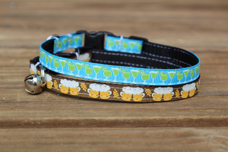 Op de afbeelding: Een bruin en blauwe kattenhalsband met een bel. De bruine halsband heeft een patroon van bierglazen en de blauwe halsband heeft een patroon van groene cocktailglazen.