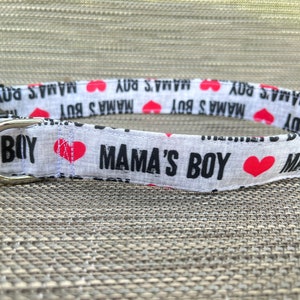 May include: A white fabric pet collar featuring a repeating black text pattern that reads MAMA'S BOY interspersed with small red heart icons. The collar includes a black plastic side-release buckle and a silver-toned metal D-ring for leash attachment.