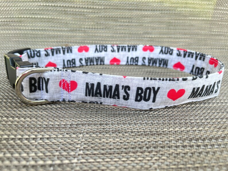 May include: A white fabric pet collar featuring a repeating black text pattern that reads MAMA'S BOY interspersed with small red heart icons. The collar includes a black plastic side-release buckle and a silver-toned metal D-ring for leash attachment.