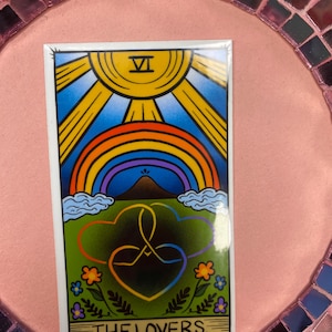 May include: A rectangular sticker featuring a tarot card design. The card depicts a sun, rainbow, and two intertwined hearts over a green field. The text "THE LOVERS" is at the bottom. The background is a pink surface with a mosaic border.