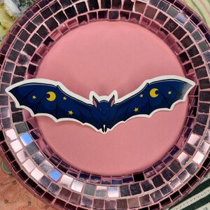 May include: A dark blue bat sticker with yellow crescent moons and stars, centered on a pink plate. The plate has a mosaic border with pink and silver tiles. The background includes floral and striped fabric.