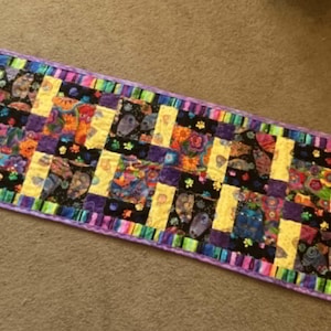 May include: A rectangular, handmade quilt with a colorful patchwork design. The quilt features a mix of yellow, purple, black, and multi-colored fabrics. The edges are bordered with a striped pattern in various colors, and the quilt is approximately 30 inches long.