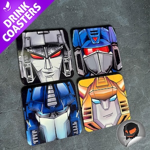 May include: Set of four square drink coasters featuring stylized robot faces in shades of gray, blue, and yellow. The coasters have a glossy finish and the words "DRINK COASTERS" are printed on a purple banner.
