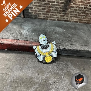 May include: A soft enamel pin featuring a stylized robot with a gold chain and pendant. The pin is primarily silver, black, and gold, with green eyes. The pin is on a gray surface with the words "SOFT ENAMEL PIN" in a banner.