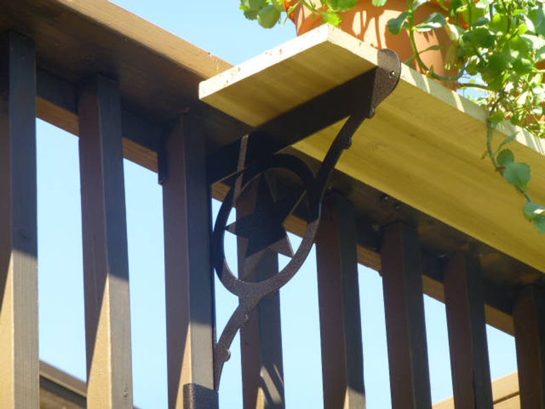 May include: A black metal shelf bracket with a decorative star design is attached to a wooden deck railing. The bracket supports a wooden shelf.