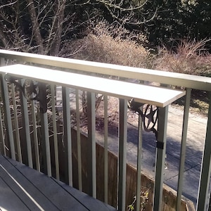 May include: A wooden shelf attached to a railing on a deck. The shelf is supported by two metal brackets. The shelf is made of light wood and the brackets are black metal.